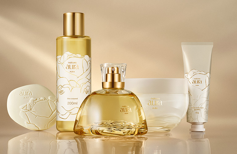Fondo bege con luz suave. Cinco productos de la línea Natura Aura Alba: frasco spray, perfume, hidratante, jabón y crema. Envases blancos y dorados.